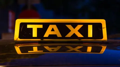 A taxi sign with illuminated yellow letters reflected on the roof of a taxi.