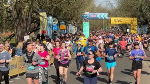 Hundreds of people are running towards the camera along a tree-lined road as part of a half marathon. There is a large sign in the middle of the road which says Cambridge. There is a mix of male and female runners and those in the foreground of the image appear to be looking around at the crowd. 