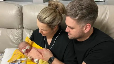 Russo family Bryony and Daniel Russo, both wearing black, sit on a cream sofa while cradling Emmy, who has tubes attached to her and is lying on a yellow blanket.