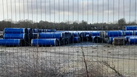 Dozens of large blue pipes stored on an empty area of industrial land with a fence in front of it.