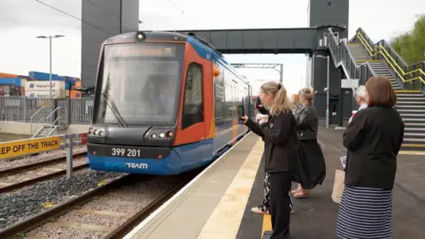 People stand on a tram platform taking photos of a blue red and orange tram