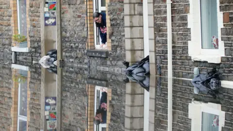 PA Media Flood water on Hunter Street in Briton Ferry, Neath Port Talbot, on Monday. A householder looks out of the window onto a flooded street from her upstairs window and she is reflected in the water below.