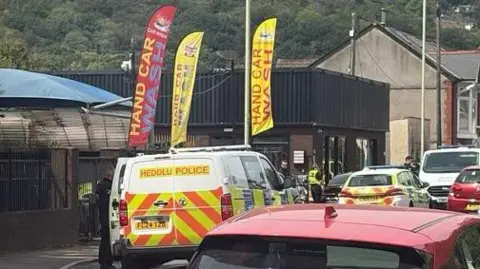 A police van and police car parked outside a car wash. There are officers also stood outside in uniform.