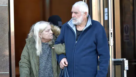 Barbara and Douglas Daniel leaving Glasgow Sheriff Court. Barbara is on the left and is wearing a large green jacket over a grey top. She has white hair. Douglas is on the right and has white hair and a beard. He is wearing a blue fleece.