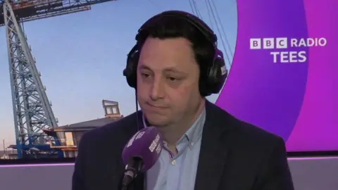 Mayor Ben Houchen is sat in the BBC Tees studio with a purple microphone in front of him. The BBC Radio Tees logo can be seen over his right shoulder, which includes a picture of the Transporter Bridge.
