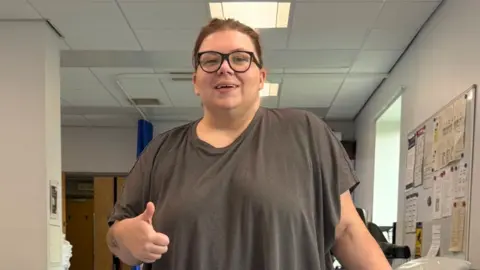 Natalia is at a physio rehab centre, stood between parallel bars to help her walk. Her left leg is in a metal cage. She makes a thumbs up pose to the camera. 