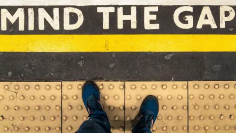 Mind the Gap sign shot taken vertically so you can see the sign on the platform and someone wearing trainers standing behind the yellow line on the bobbled paved section. Above the yellow line there is a Mind the Gap sign painted in white bold capital letters.