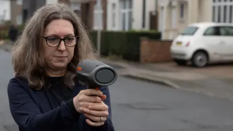 A middle-aged woman with long, greying hair, standing in a road, holding up a speeding gun