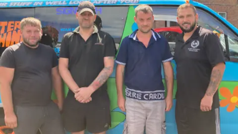 Graham Wilde, Matt McGinley, Steven Buckley and Scott Boraston (left to right) are stood in front of their van. They're wearing T-shirts and shorts and all have short beards.