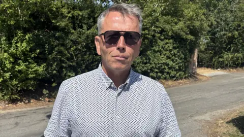 Luke Deal/BBC A man stands outside and smiles at the camera. He has short grey hair, sunglasses and is wearing a white shirt with blue markings on it. There is a road behind him, as well as a tall hedge lining the road on the opposite side. 