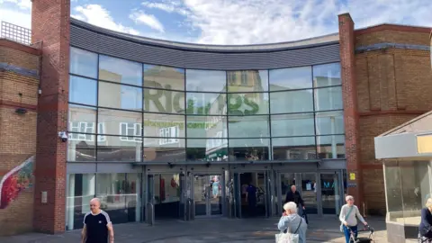 A brick and glass frontage of a shopping centre with people walking outside