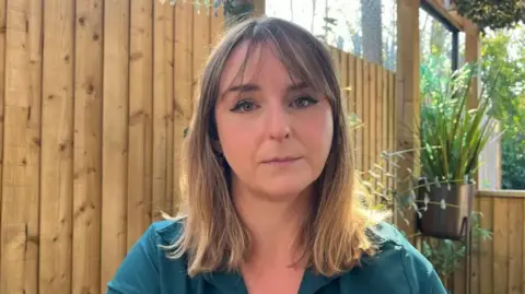 BBC Crystal has long, brown shoulder-length hair and is wearing a green top. She is sitting outside by a wooden fence with plants behind her. The sun is shining.