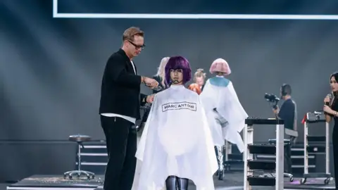 Bruno Marc Giamattei Bruno Marc Giamattei is cutting a purple wig on a mannequin. He has glasses and a black blazer on.