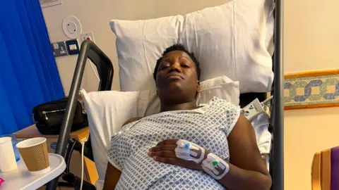Abigail Chirwa A woman lying on a hospital bed wearing a gown. She has her eyes closed. She has a cannula in her hand.