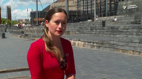 BBC A woman in a red dress with long, dark hair sits outside the Senedd in Cardiff Bay.