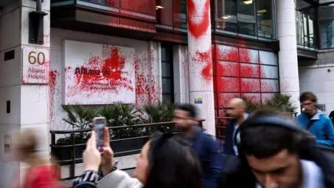 Reuters White-fronted office building in London with pillars between the windows. Much red paint is visible on the frontage of the building - including on the Allianz sign.  People are walking past and one is taking a picture on a mobile phone.