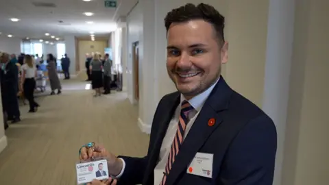 An image of a Joshua Roberts standing in a corridor, smiling and holding up his councillor photo ID. In the background there are other people milling about.