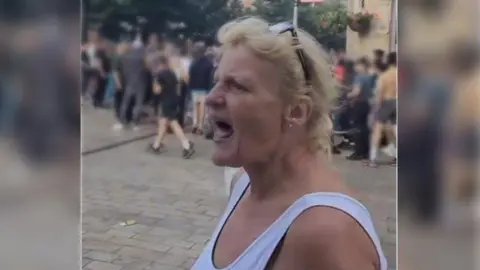 A woman with blond hair and glasses on her head, dressed in a white vest, is filmed while screaming