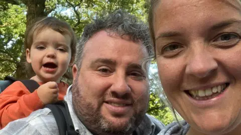 Indie (left), Andy Inkpen (centre) and Jody Peters smiling and looking at the camera. They appear to be in the woods and Indie is in a carrier on Andy's back.