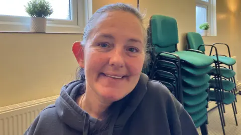 A woman in her forties with greying hair in a ponytail, wearing a grey hoodie and sitting on a chair