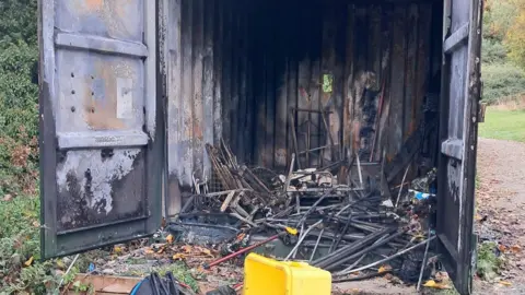 Higham Ferrers Riverside Angling Association A large storage container, with its doors open, with burnt and damaged items all around it, a yellow box at the front, and burnt and damaged items on the ground.