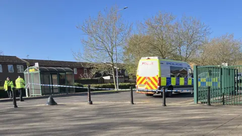 Shariqua Ahmed/BBC A police scene remains in place near a bus stop. Two officers in high-vis vests are standing in the road, and a police van and car are parked on the street. Police tape is in place.