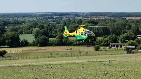 A yellow and green helicopter hovering a short distance from the ground as it prepares to land, in the middle of a field, with a several fences in the distance in front of two small buildings and a large number of trees leading to the skyline