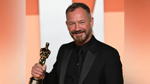 Lee Sandales has short dark hair and a dark beard. He is wearing a black suit with a black shirt and black tie and is holding an Academy Award in his right hand.