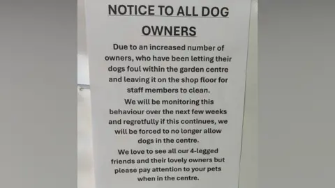 A white sign with black writing which has been displayed in the garden centre. The top of the sign reads 'Notice to all dog owners'.