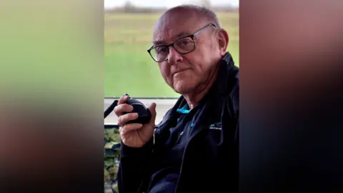 Roger Smith Roger Smith looks at the camera and smiles. He holds a small black speaker in his hand which is raised toward his ear. He is bald and is wearing glasses along with a navy jacket and top.