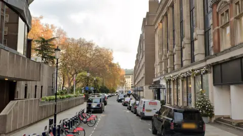 A Google Maps street view screen grab of Seville Street. Black cabs are parked on the left and cars and a white van are on the right. What looks like a residential building is to the left and shops on the right.