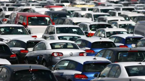 A large amount of cars all lined up. All you can see are cars parked very closely together. All number plates are not visible and all the cars appear to be parked with nobody in them.