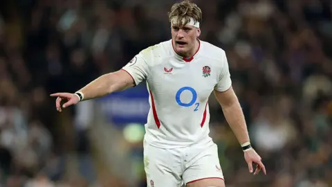 Getty Images Alex Cole playing for England. He is wearing a white England rugby kit which is slightly dirty with mud and grass. He is looking to the side and pointing.