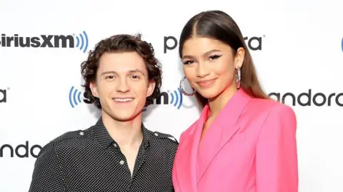 Getty Tom Holland and Zendaya stand on a red carpet. Tom wears a black and white spotty shirt and Zendaya wears a bright pink suit, silver hoops and long brown hair 
