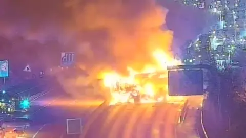 CCTV footage of a large fire on a motorway. Large flames can be seen and one fire engine.