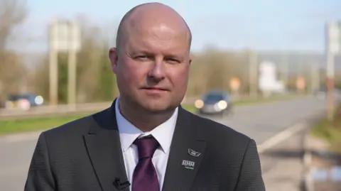 Geraint Evans, a bald man in a dark grey suit and purple tie, is looking into the camera. It's a sunny day, and he is standing at the side of a dual carriageway in Pencoed, which is blurred in the background. 