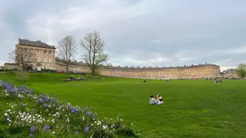 An image of the Royal Crescent lawn where a handful of people are sitting and enjoying a picnic. The iconic row of Grade I terrace houses are in the background.