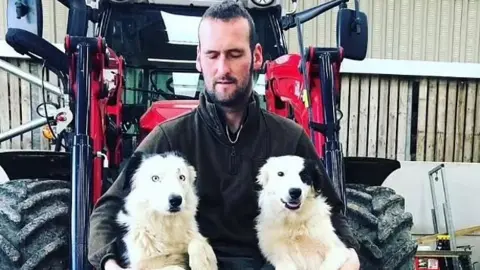 Ricky Rennie Ricky Rennie, wearing black trousers, jumper and boots, sits on a digger outside a barn with two sheep dogs leaning on his legs.