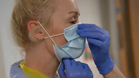 Getty Images A nurse puts on a surgical face masks, she wears blue plastic gloves, has blonde hair and a double ear piercing