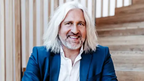 Christian Ege Head shot of Christian Ege with shoulder-length grey hair and beard sitting on some wooden steps. He is wearing a blue suit with a white shirt with the top buttons unopened.