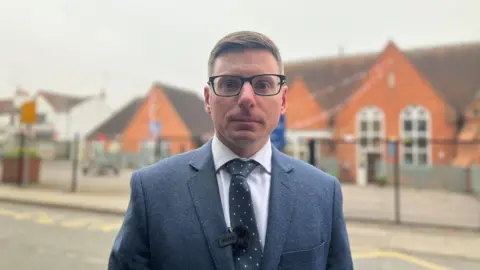 Howard is wearing a blue blazer with a blue and white polka dot tie and is behind the school