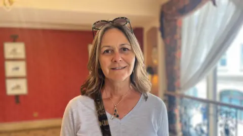 Kim Cisna is standing in a hotel reception room with red walls and a large window. She is wearing sunglasses on her head, with a grey top. She is smiling into the camera.