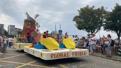 A float vehicle covered in flowers on a road, made to look like a windmill and two giant clog shoes. There are children in costumes sat inside the shoes and stood by the windmill. The front of the float says 'FLORAL FRISIA'. On either side of the road, there are small crowds of people watching the float go past. Some people are taking photos. 