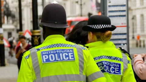 A photograph of the back of one male and one female Metropolitan police officers, walking through the streets of London