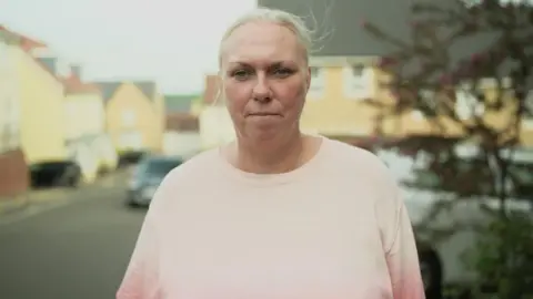 A woman with blond hair bound to look at the camera. She does not smile. She wears the pale pink bird and stands on a residential street, which is not clear