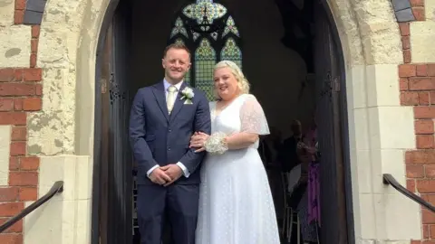Dudley Metropolitan Borough Council A couple stand in the arch of a church on their wedding day. On the left the groom is wearing a dark blue suit with a cream tie. The bride has short blonde hair and is wearing a flowy white polka dot dress. They are smiling and looking down at the camera. 