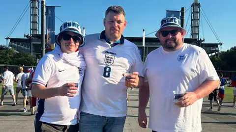 Andy Taylor Three Lions superfan Andy Taylor with his friends. He is wearing an England shirt and bucket hat, and has a St George's flag with him. He is pictured with two friends, who are both wearing England shirts. Behind them is a German football stadium.