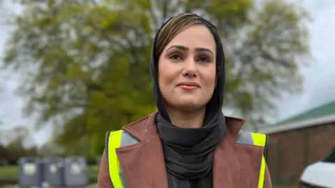 A young woman in head scarf and hi viz vest looks towards the reader.