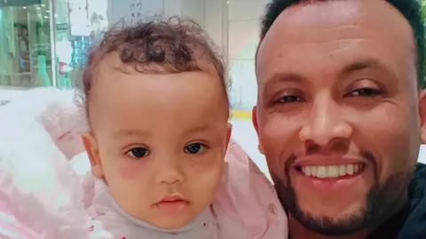 Nestere and his young daughter look directly at the camera smiling. The little girl is wearing a pink coat.