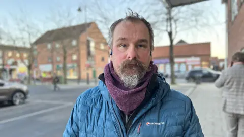 A man with a greying beard stands on a street. He is wearing a blue jacket and a purple snood. Trees and brick buildings are in the background.